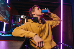 Portrait of young professional cyber sportsman sitting at gaming desk at home and drinking caffeine energy drink to concentrate on online video shooter game at night. Neon coloured room. Cyber sport