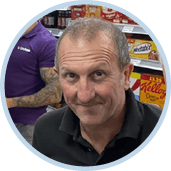 A man is smiling in front of a store with a shelf full of food. Description generated by AI