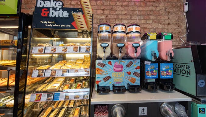 A bakery display case filled with various pastries is next to a counter with colorful frozen drink dispensers labeled with different flavors. Description generated by AI