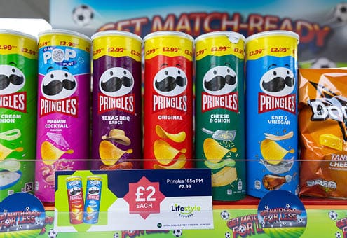 A row of six colorful Pringles cans with different flavors is displayed on a shelf with a price sign in front. Description generated by AI