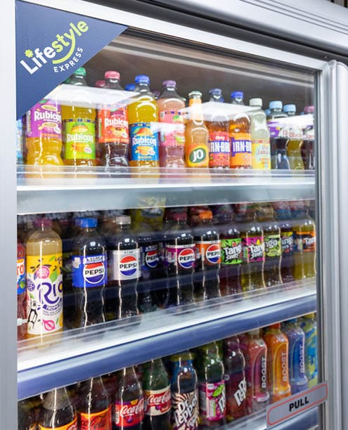 A refrigerated display case filled with various bottled soft drinks including Pepsi, Coca-Cola, Rubicon, and other colorful beverages arranged on three shelves. Description generated by AI
