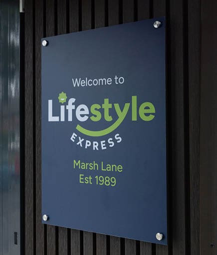A dark blue sign mounted on a wooden wall reads welcome to Lifestyle Express Marsh Lane Est 1989 with the word Lifestyle highlighted in white and green. Description generated by AI