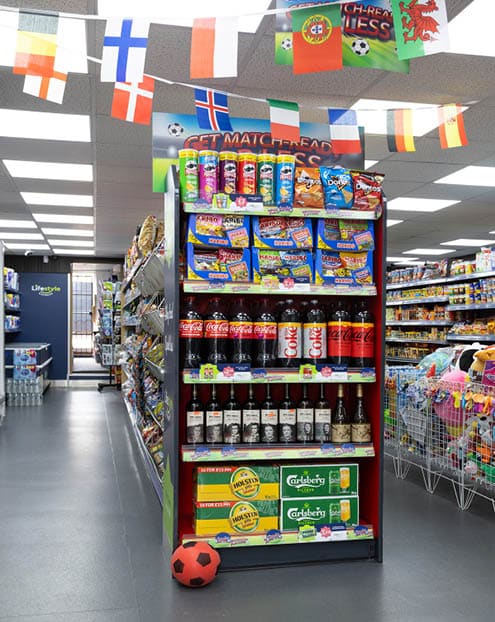 A supermarket aisle displays shelves stocked with snacks, sodas, and beer with international flags hanging from the ceiling and a small red and black soccer ball on the floor. Description generated by AI