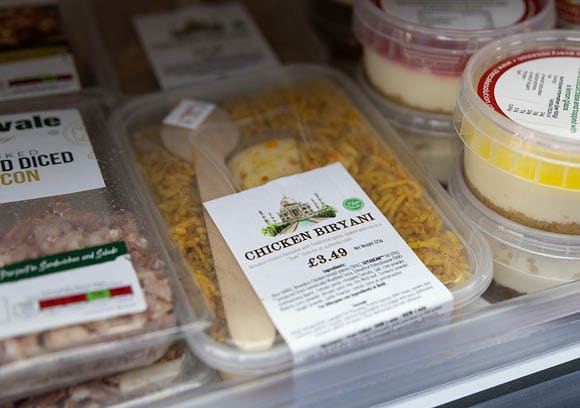 A packaged ready-to-eat chicken biryani meal with a wooden fork inside a refrigerator next to other packaged food items. Description generated by AI