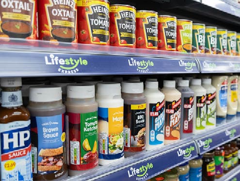 Shelves in a store display various bottles and cans of sauces, soups, and condiments with colorful labels and price tags. Description generated by AI
