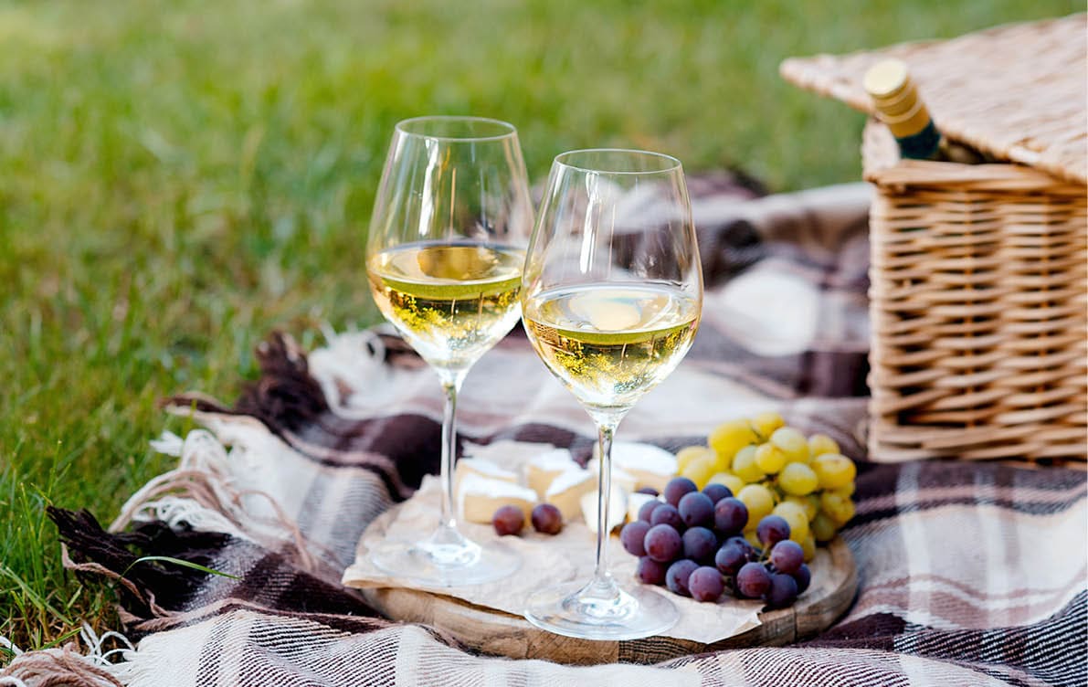 Two glasses of white wine, grapes, cheese, and nuts are arranged on a blanket next to a wicker picnic basket on grass. Description generated by AI