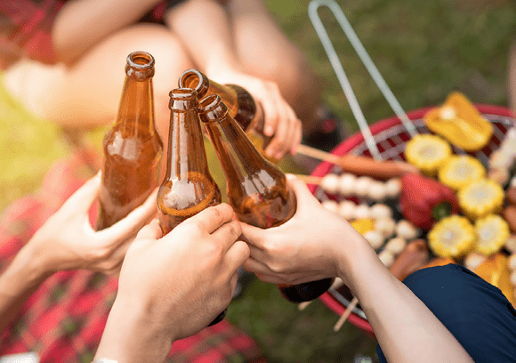 People sitting outdoors clinking brown glass bottles together near a grill with skewers of food and corn. Description generated by AI