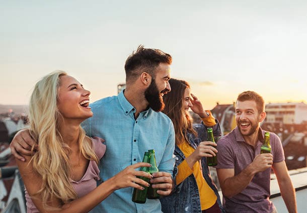 Four people stand close together on a rooftop at sunset, holding green bottles and smiling while enjoying a casual social moment. Description generated by AI