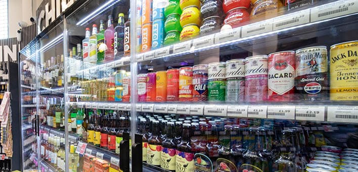 A refrigerated display case filled with various colorful canned and bottled beverages arranged on multiple shelves in a store. Description generated by AI