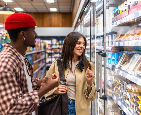 Two adults stand in a grocery store aisle looking at refrigerated food items while one gestures and the other holds a large black bag. Description generated by AI