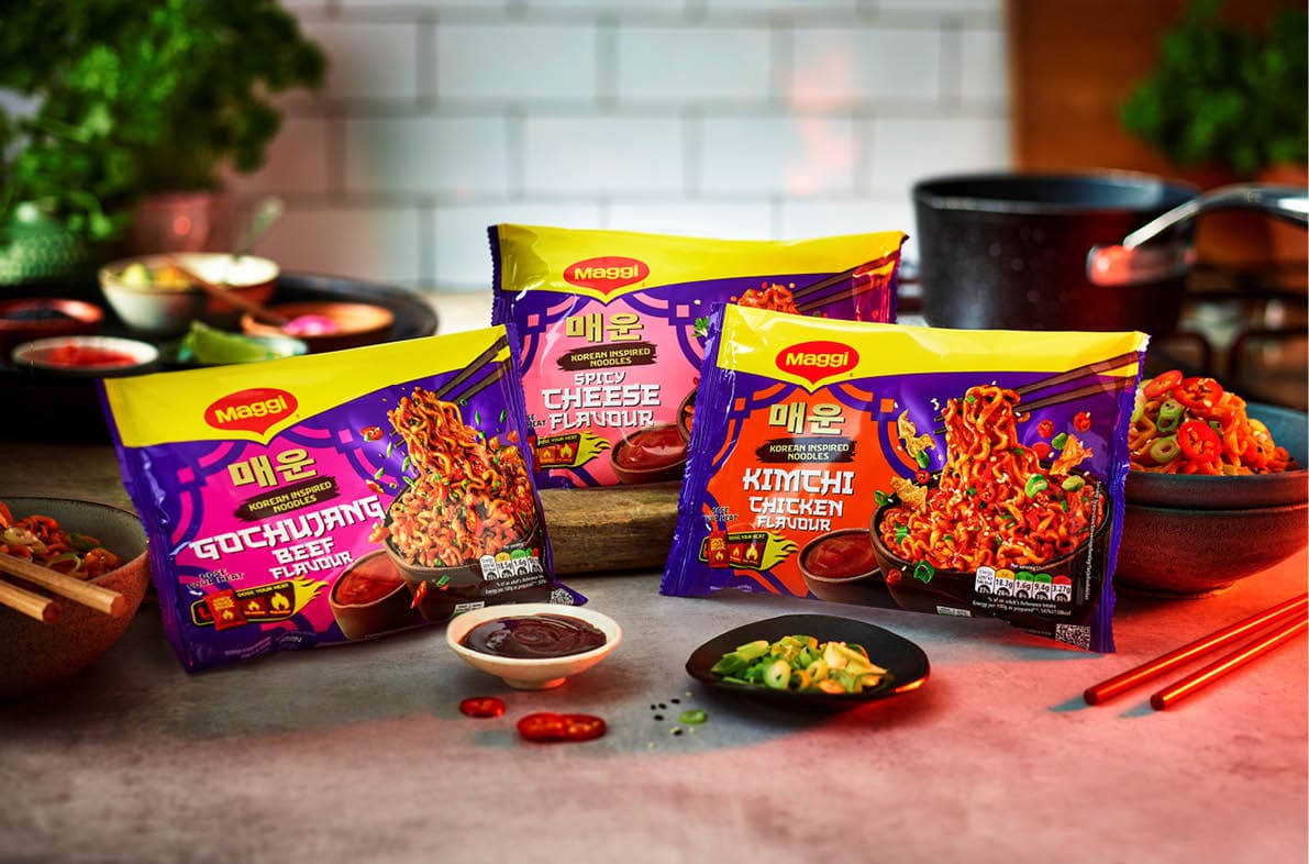 Three packets of Maggi spicy instant noodles with gochujang, cheese, and kimchi chicken flavors are displayed on a kitchen counter with bowls of sauce, chopped vegetables, and chopsticks nearby. Description generated by AI
