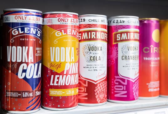 Cans of flavored vodka drinks in various bright colors are lined up on a shelf with promotional price tags. Description generated by AI