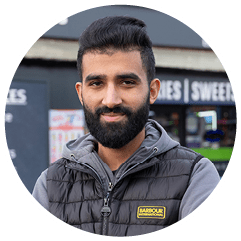 A person with styled hair wearing a black Barbour International vest over a gray hoodie stands outdoors in front of a storefront. Description generated by AI