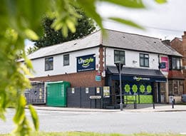 A corner store named Lifestyle Express with a white upper facade and brick lower walls is situated at an intersection with green foliage partially framing the view. Description generated by AI