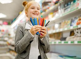 A smiling child with hair in a bun wearing a gray blazer and white shirt is holding a colorful bundle of markers in a store aisle. Description generated by AI