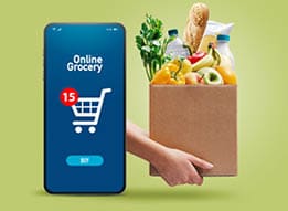 A person holds a box filled with groceries next to a smartphone screen displaying an online grocery shopping app with a shopping cart icon and a notification. Description generated by AI