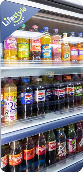 A refrigerated display case filled with various bottled soft drinks including Rubicon, Pepsi, Coca-Cola, Dr Pepper, and other brands. Description generated by AI
