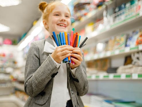 Person wearing a gray blazer and white shirt holding a colorful assortment of markers inside a store with shelves of various items in the background. Description generated by AI