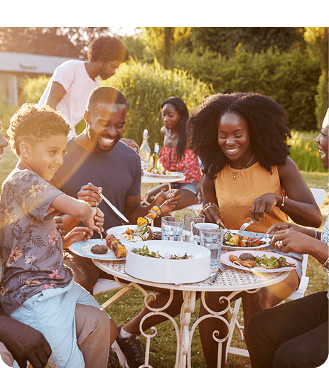 A group of people with curly and natural hairstyles are happily enjoying a meal together outdoors at a round table with plates of food and drinks in a sunny garden setting. Description generated by AI