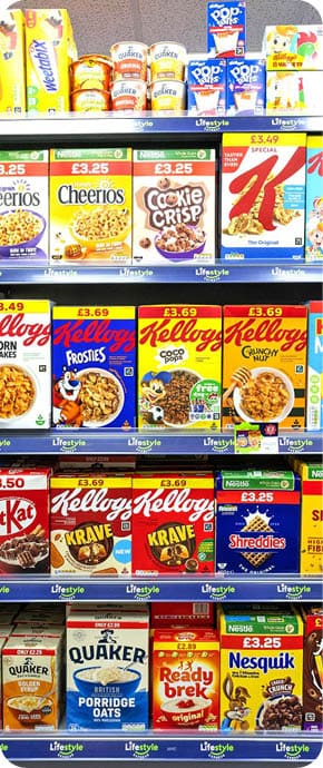 A supermarket shelf displays various breakfast cereals and related products including Cheerios, Cookie Crisp, Special K, Kellogg's Frosties, Krave, Shreddies, Quaker porridge, Ready Brek, and Nesquik in colorful packaging. Description generated by AI