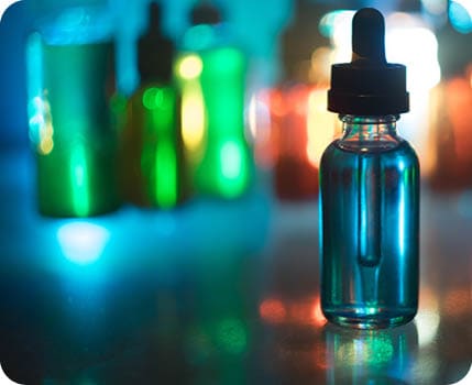 A clear glass dropper bottle filled with blue liquid is in sharp focus on a reflective surface with several blurred colorful bottles in the background. Description generated by AI