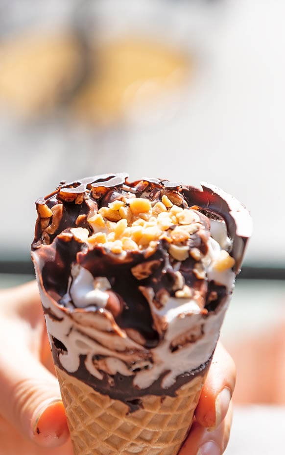 Hand holding one white vanilla chocolate swirl ice cream gelato cone with bokeh background on hot summer day and peanuts topping