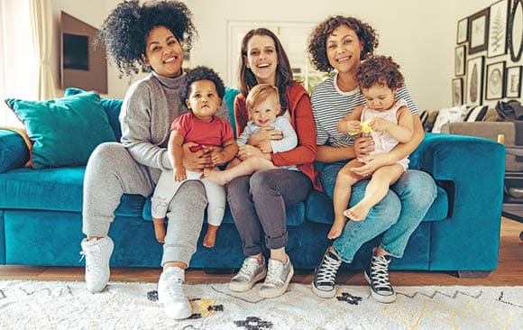 Multi-ethnic mommies sitting with children. Young mothers with babies sitting on sofa and looking at camera.