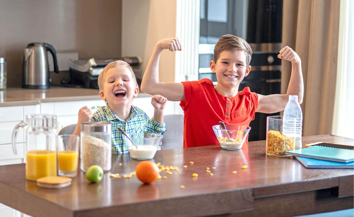 Smiling kid and his happy younger sibling flexing their muscles at the table during the morning meal
