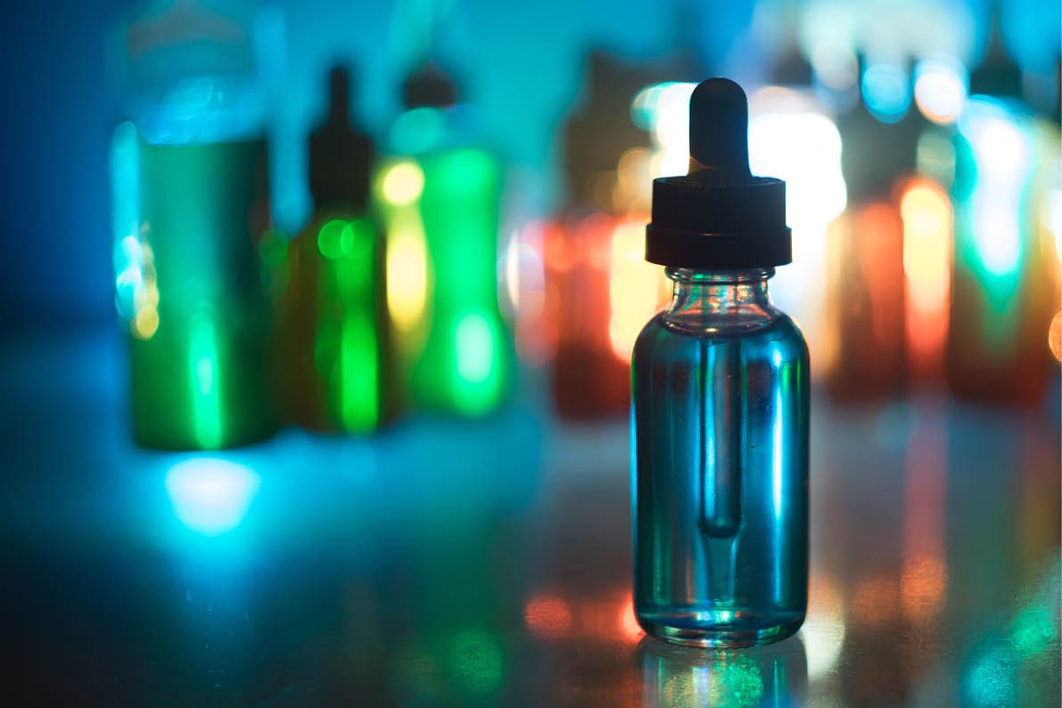 A clear dropper bottle filled with blue liquid is in sharp focus on a reflective surface with several blurred colorful bottles in the background. Description generated by AI