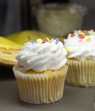Two vanilla cupcakes with white frosting and colorful round sprinkles sit on a gray surface with a blurred background. Description generated by AI