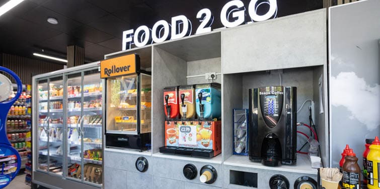 A self-service food and beverage station with refrigerated shelves stocked with drinks and snacks, a coffee machine, and condiment dispensers under a sign that reads FOOD 2 GO. Description generated by AI