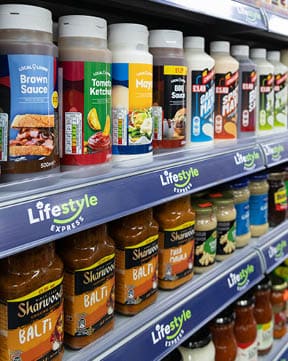 Bottles of various sauces and condiments including brown sauce, tomato ketchup, mayonnaise, BBQ sauce, and curry sauces are neatly arranged on supermarket shelves with Lifestyle Express branding. Description generated by AI