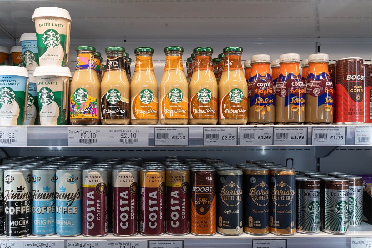 A refrigerated shelf displays various bottled and canned coffee drinks from brands including Starbucks, Costa, and Boost arranged in rows. Description generated by AI