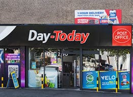 A storefront with a black sign reading “Day-Today" and a red "Post Office" logo, featuring promotional posters for grocery delivery and nicotine pouches on the windows. Description generated by AI