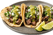 Three tacos filled with grilled meat, chopped onions, cilantro, and green salsa are served on a plate with lime wedges. Description generated by AI