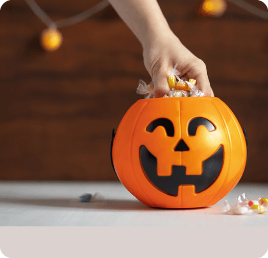 A hand is reaching into an orange plastic pumpkin bucket filled with wrapped candy on a table with blurred string lights in the background. Description generated by AI