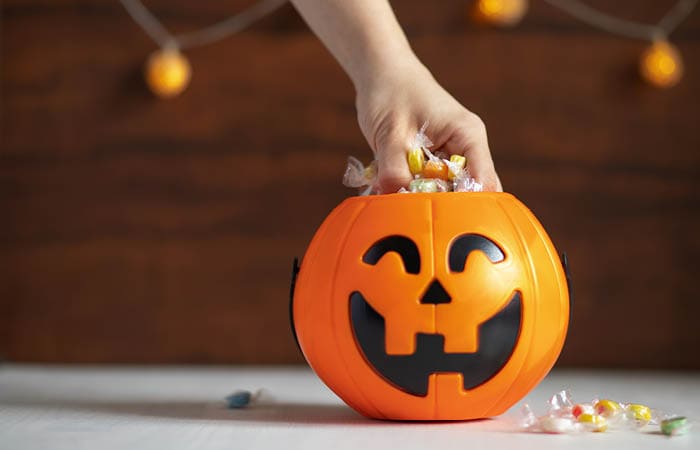 A hand is reaching into an orange pumpkin-shaped container filled with wrapped candies on a white surface with blurred string lights in the background. Description generated by AI