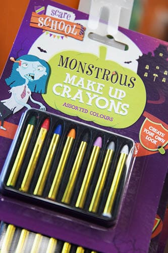 A colorful package labeled “Monstrous Make Up Crayons" features eight gold and colored crayons with a cartoon character and Halloween-themed design. Description generated by AI