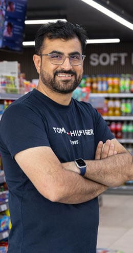 A person wearing glasses and a navy blue Tommy Hilfiger t-shirt is standing with arms crossed in a brightly lit store aisle with shelves of colorful products. Description generated by AI