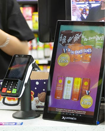 A digital display at a checkout counter shows promotional prices for various soda and flavored drink cans next to a card payment terminal and a person standing nearby. Description generated by AI