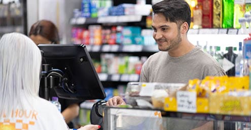 A person with short dark hair and a gray shirt is smiling and interacting with another person with long white hair at a store counter with various products and a computer screen. Description generated by AI