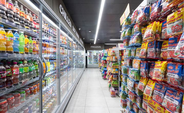 A supermarket aisle with refrigerated shelves on the left stocked with beverages and frozen items, and racks on the right filled with various packaged snacks. Description generated by AI