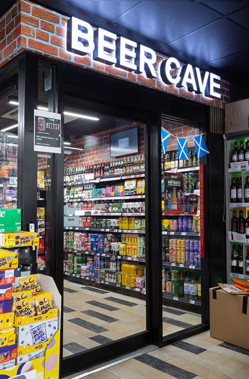 A brightly lit store entrance labeled Beer Cave displays shelves stocked with various canned and bottled beverages inside. Description generated by AI
