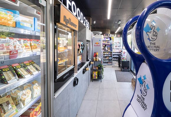 A convenience store aisle features refrigerated shelves stocked with sandwiches and snacks, a hot food dispenser labeled “Rollover," and a blue lottery ticket machine with "The National Lottery" logo. Description generated by AI