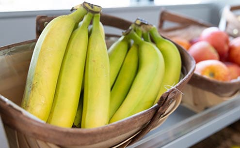 A wooden basket filled with ripe yellow bananas is placed on a shelf next to another basket containing red apples. Description generated by AI